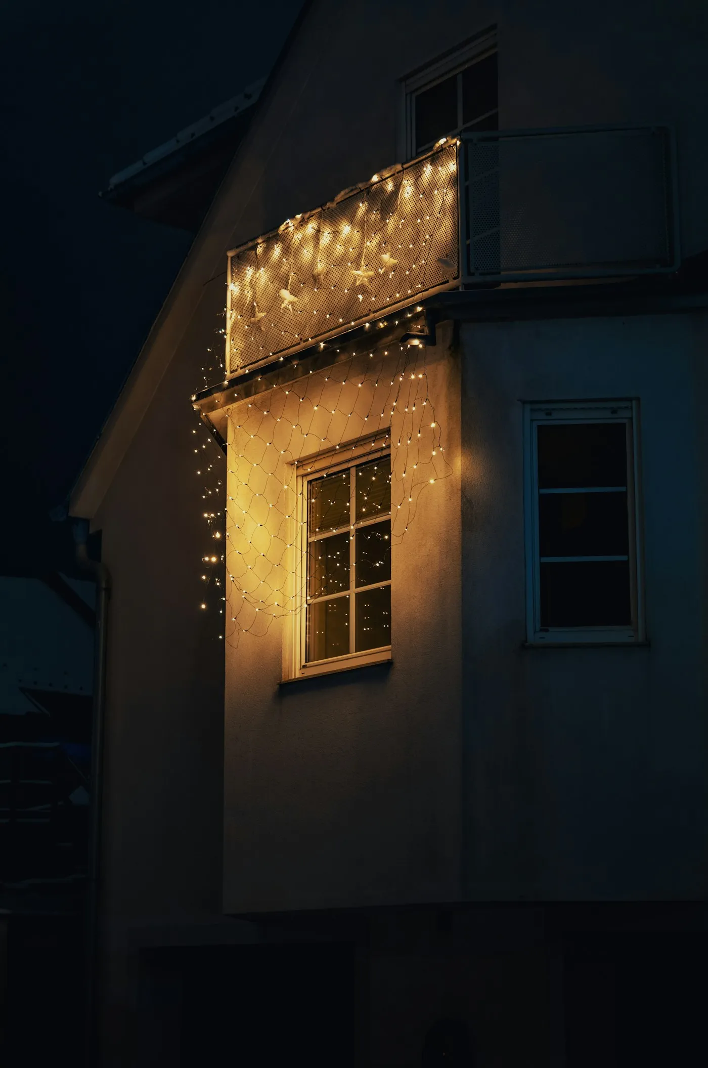 Warm string lighting running down the front of a house after dark, lighting the wall and window from above.